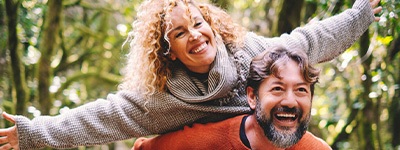 Woman with curly hair riding on back of man in orange sweater in forest