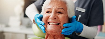 Woman in dental chair smiling with dentist’s gloved hands touching her cheeks
