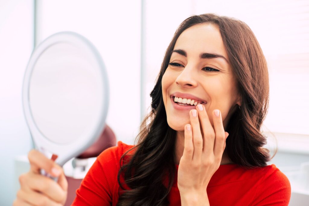 Woman in red-orange shirt touching her teeth smiling into mirror